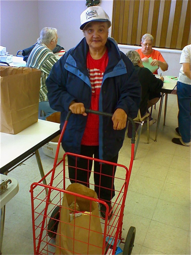 An elderly person smiles and poses with a grocery cart containing food.
