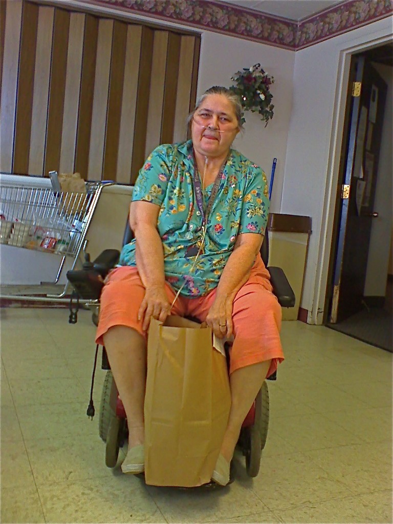A woman using a power scooter and oxygen tank poses holding a brown paper bag full of groceries.