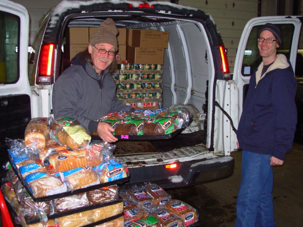 Two men smile as they unload palettes of groceries from a white truck.
