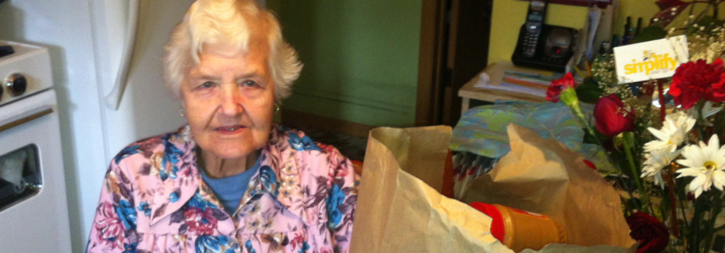 An elderly woman wearing a floral patterned shirt sits in her kitchen and softly smiles. A brown paper bag full of groceries sits on the table in front of her.