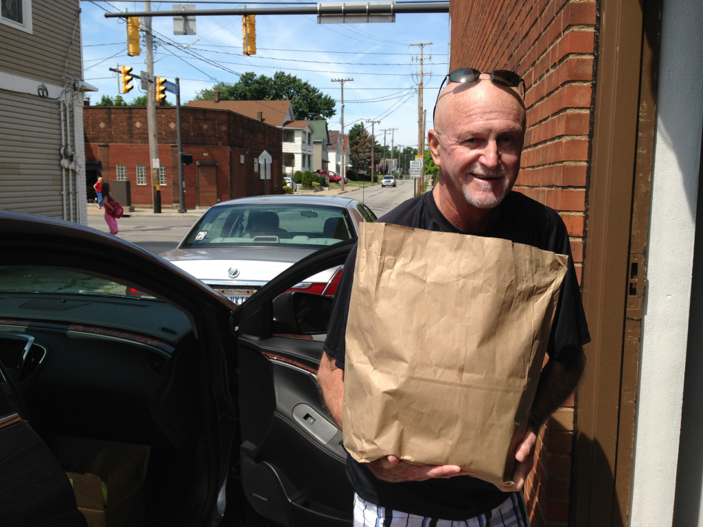 An All Faiths Pantry volunteer smiles as he holds a brown paper bag full of groceries. 