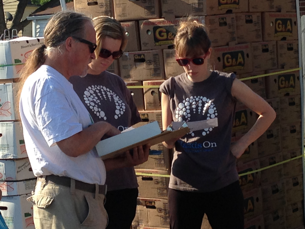 Three volunteers discuss and collaborate while standing in font of palettes stacked high with boxes of produce. A man points to a sheet of paper on a clipboard and a woman has her hands on her hips. 