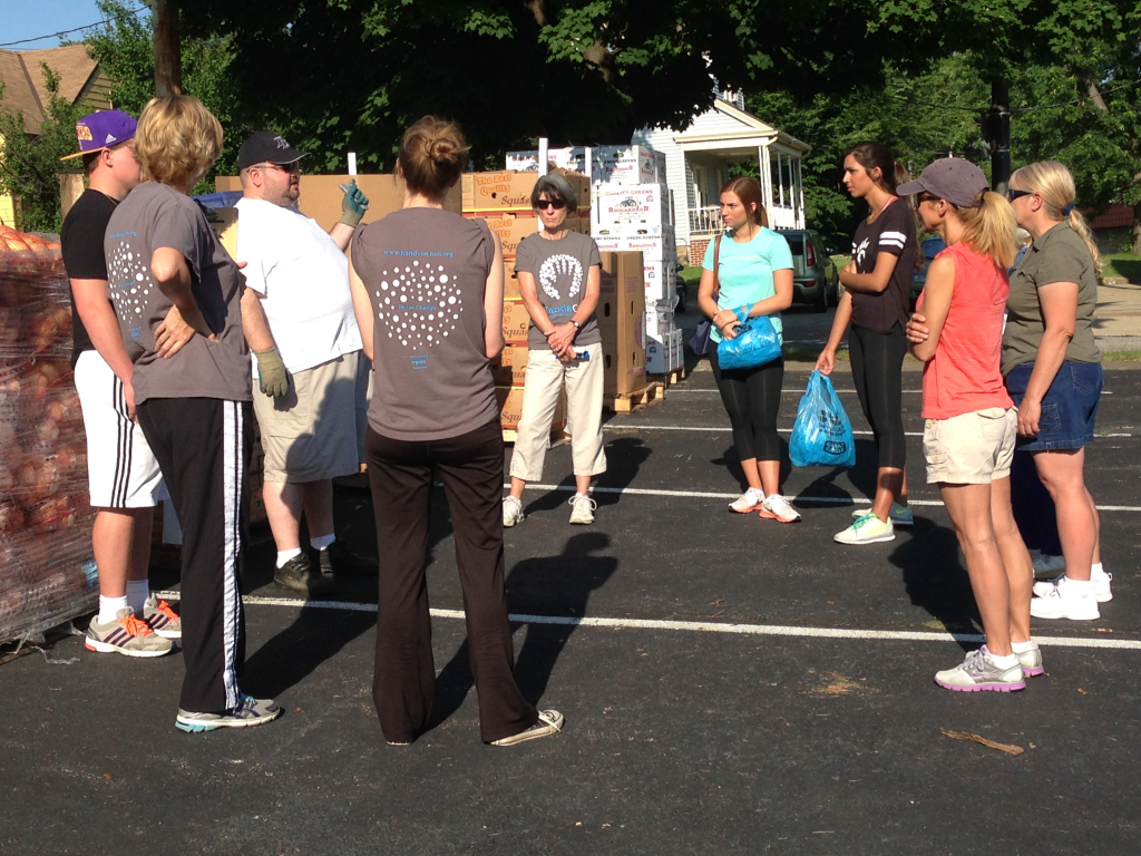 A group of nine volunteers stand in a circle near a row of palettes of food. One volunteer debriefs the group on their task. They are standing outside in a parking lot on a sunny day. 