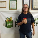 Founder John Visnauskas holds a jar of peanut butter and smiles. He is standing in front of a wall where various awards and an All Faiths Pantry banner are hung.