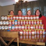 Two men and two women smile widely. They are standing behind a stack of jars of peanut butter than nearly obscured their faces.