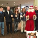 Six men and women, including a man dressed as Santa Claus, hold jars of peanut butter and pose in front of a collection box.