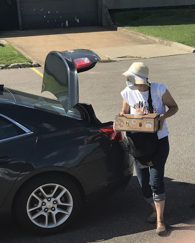 A woman wearing a sun hat and face mask loads a box of groceries into the trunk of a car.