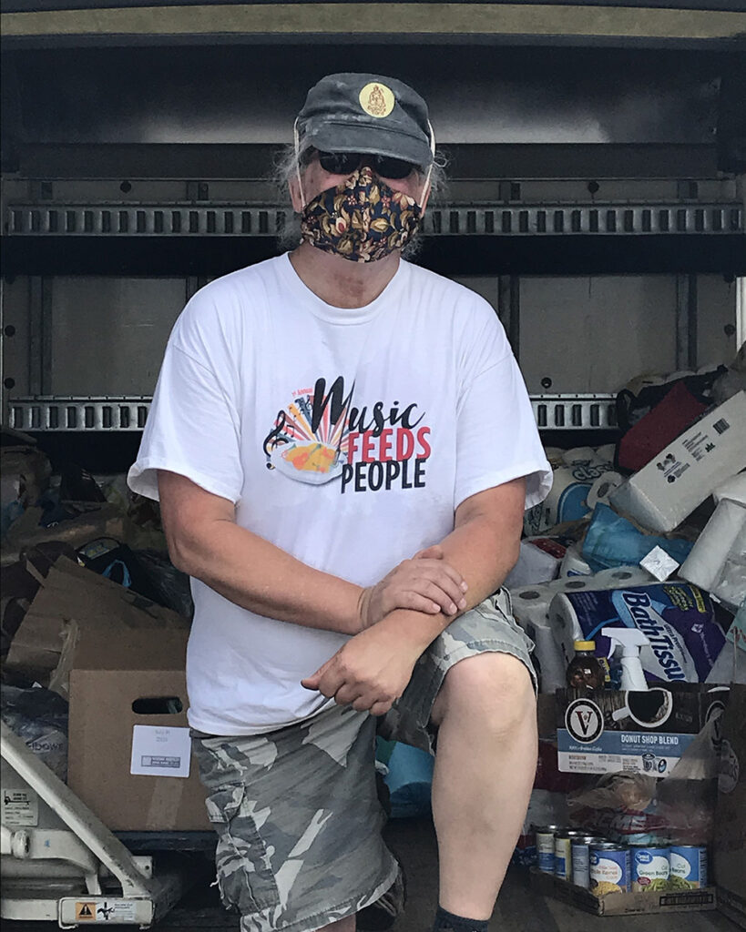 A man wears a mask and poses kneeling on one knee. He is inside of the back of a box truck full of groceries.