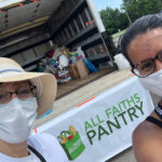Two women wearing face masks pose in front of a banner with the All Faiths Pantry logo.