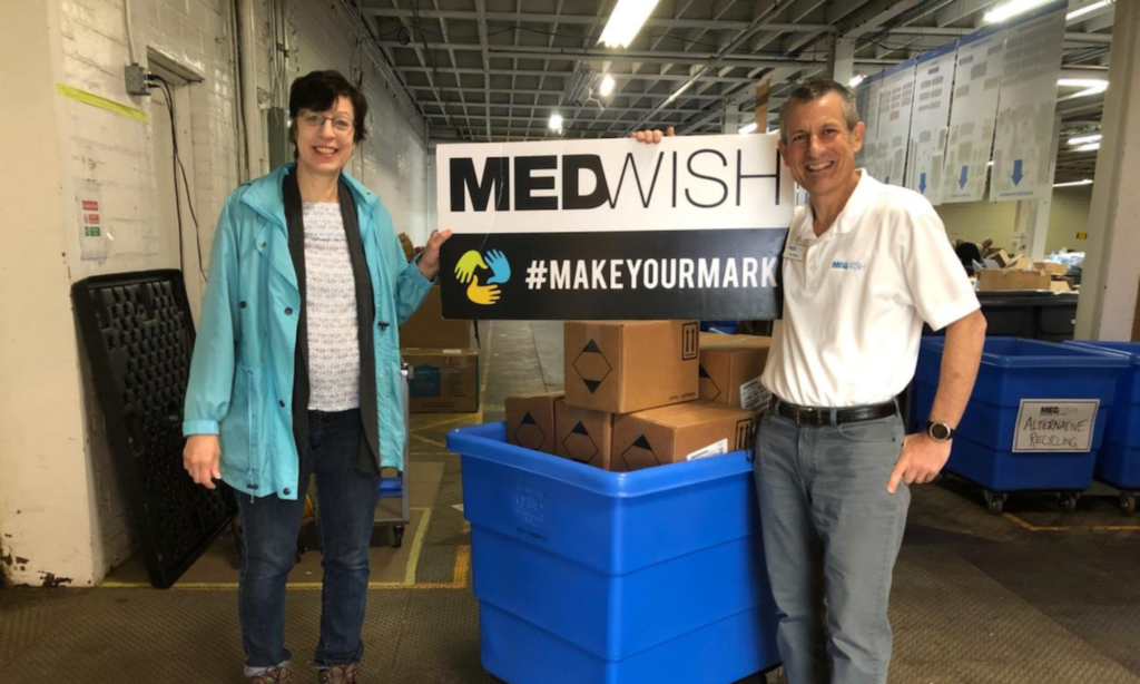 A man and a woman stand in front of a bin full of cardboard boxes. They smile and hold a banner that reads: "MedWish #make your mark"