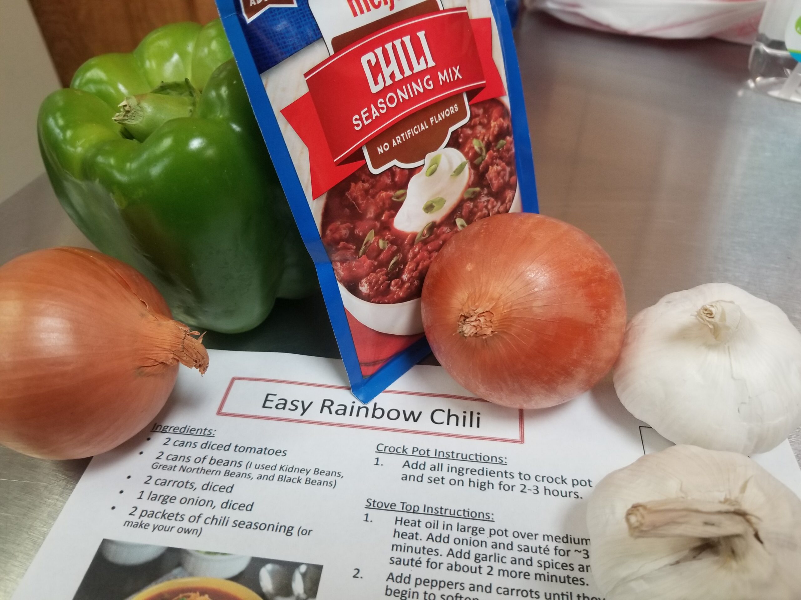 A piece of paper with a recipe for "Easy Rainbow Chili" sits on a stainless steel table, surrounded by some of the ingredients to make the dish (chili seasoning, onion, garlic, and a green bell pepper.