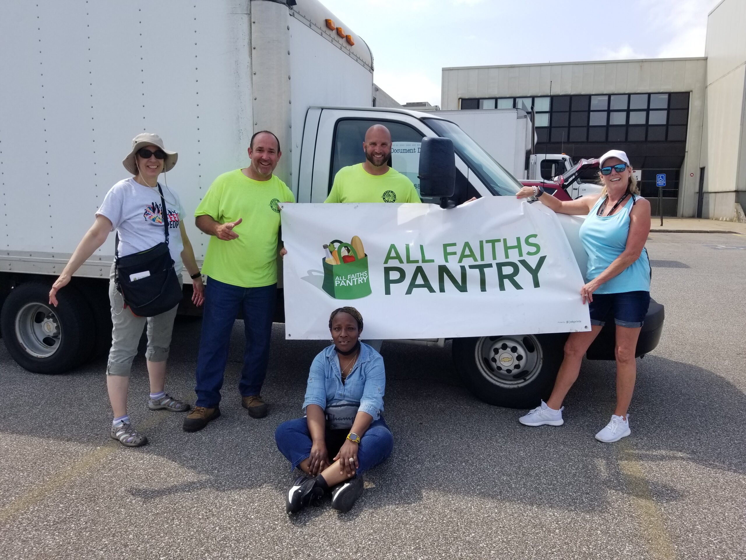 Four people stand and hold an All Faiths Pantry banner in front of a large truck. One woman sits in front on the ground. They are in an industrial parking lot.